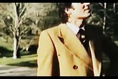 Person in a tan double-breasted coat looking up among trees in a park, 16 anos 7.
