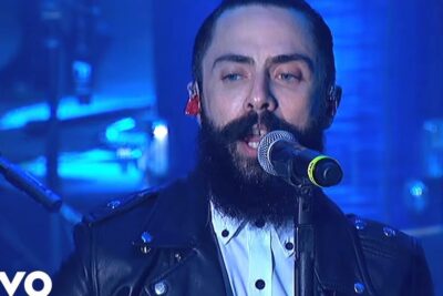 Bearded vocalist singing into microphone on blue-lit stage with 16 anos 8