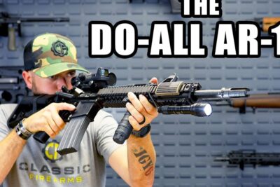 Person aiming a tactical AR-style carbine with optic, mounted flashlight and curved magazine, 16 ar 1