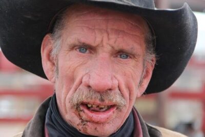 Close-up portrait of a weathered cowboy in a black hat and scarf with blue eyes and a rough beard, evoking the spirit of 4 maneders.