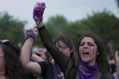 Young women with purple bandanas and painted faces raising fists at an outdoor protest 8 marzo 1