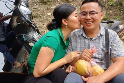 Woman kissing a smiling man holding a bag of fruit beside a parked scooter, a beskytte affectionate roadside scene.