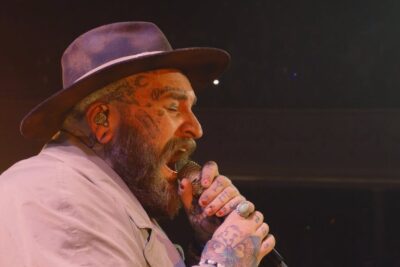 Tattooed bearded singer in a hat singing into a microphone with a lose, intense expression