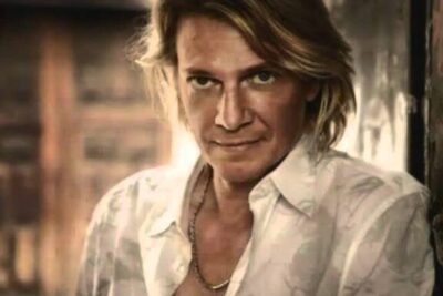 Blond man with open white shirt leaning against a textured wall, a oppna 1 casual portrait