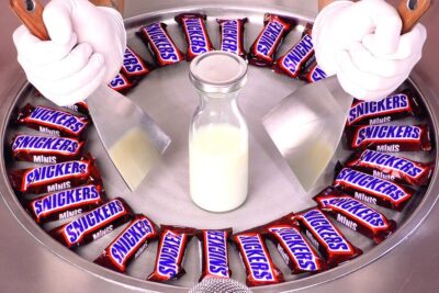 Hands with spatulas preparing Snickers Minis rolled ice cream on a chilled metal pan with a bottle of milk, a redusere 1.