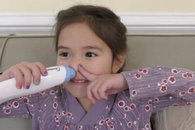 Young smiling child on a sofa using an electric nasal aspirator for gentle at-home relief — Child Using Nasal Aspirator on Sofa — a redusere 2 Product Shot.