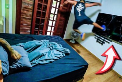 Person mid-air approaching wall-mounted a televisao with a cracked screen in a bedroom.