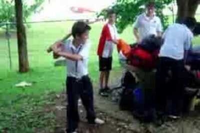 Group of students in a grassy park with one boy swinging a stick near a bench a vare 3
