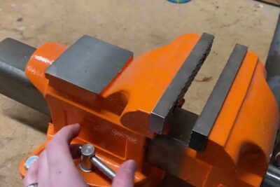 Close-up of a bright orange bench tool showing a vise with serrated metal jaws on a wooden workbench.