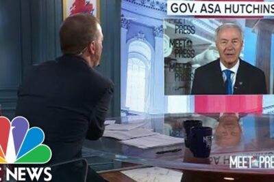 Television studio with host facing a remote guest on a large screen, NBC News branding and reflective desk, abort ar 1.