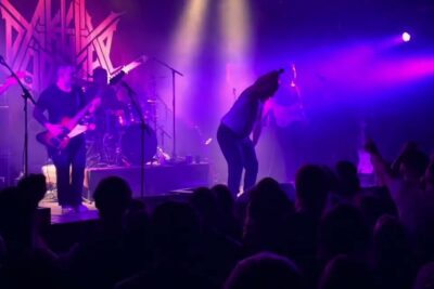 Live rock band performing under purple stage lights with a cheering crowd, aktiv dodshjelp 1.