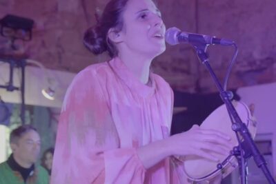 Female vocalist playing a tambourine and singing into a microphone under purple stage lights, alem disso 48.