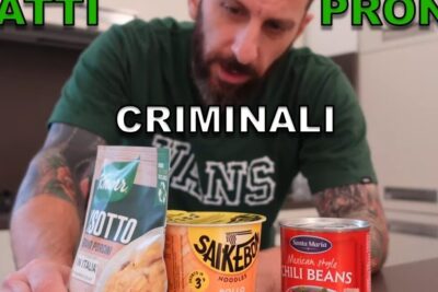 A tattooed man in a kitchen presents three packaged ready meals — a bag of risotto, a cup noodle and a can of chili beans — highlighting alimenti precotti 3