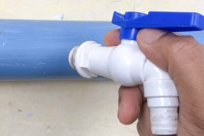 Hand holding a white PVC faucet with a blue handle connected to a blue PVC pipe, illustrating alternativa metoder.