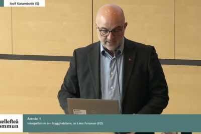 Person speaking at a lectern with a laptop and microphone during a municipal meeting about andringen tidsplanen.