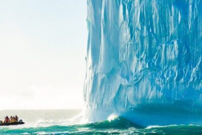A towering blue iceberg looms beside a small inflatable boat with passengers in cold-weather gear, antartida continente 1.