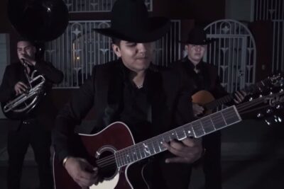 Three musicians in black suits and cowboy hats playing acoustic guitars and a sousaphone in a dimly lit street, aquellos oponen 1.