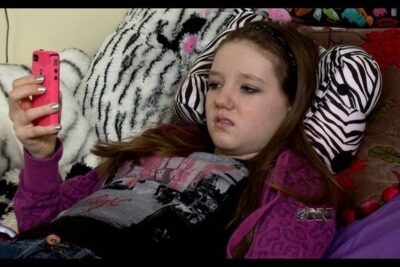 Young person reclining on a bed holding a pink smartphone with a contemplative expression for ar eutanasi coverage.
