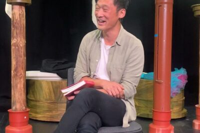 Seated person smiling on stage holding a red book during an argumenter imod 1 session.