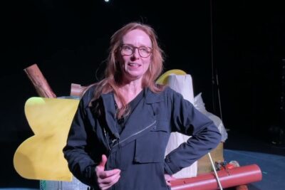 Woman on stage in glasses speaking near colorful set pieces, argumenter imod 2.