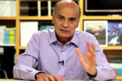 Elderly man in a purple shirt gesturing while speaking about argumentos homossexualidade in a study-like setting.