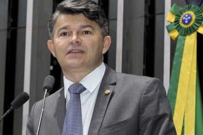 Man in a suit speaking at a podium with a microphone beside a green and yellow ceremonial ribbon, presenting argumentos propostas.
