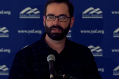 Male speaker with glasses at a microphone in front of a logo backdrop illustrating arguments abortion.
