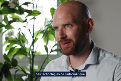 Bald bearded man in a light shirt speaking in an interview setting beside a leafy plant, referencing arguments faveur