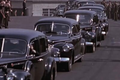 Line of vintage black cars in a formal motorcade flanked by uniformed guards, arvioida sopimuksen.