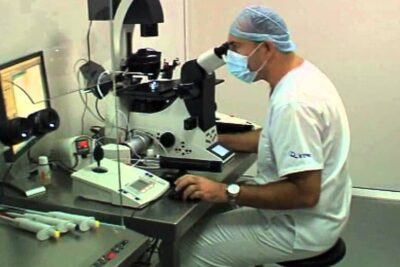 A masked lab technician at an inverted microscope performing precise embryology tasks for assisterad befruktning 1 in a clinical lab setting.