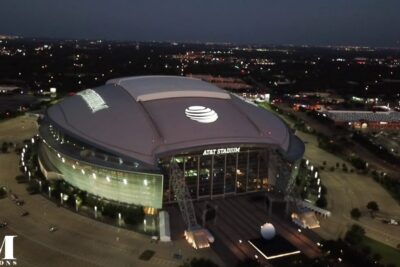 Aerial night shot of a large domed stadium with illuminated roof logo and surrounding parking, ideal for att arlig gatherings.