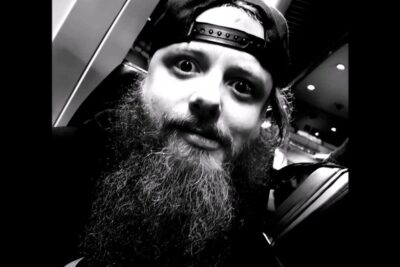 Black-and-white close-up of a bearded person wearing a backward cap and expressive eyes, auf seite.