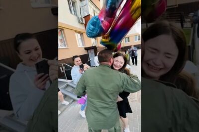 Young woman tearfully embraces a person in a green uniform holding large rainbow foil balloons while friends record the reunion, auttaa meita 12