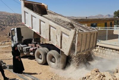 Dump truck tipping and unloading gravel and crushed stone at a rural construction site with two onlookers auttaa meita 16