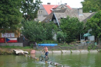 Cyclist balancing a bicycle across a narrow wooden pier over a calm river, auttaa meita 2.