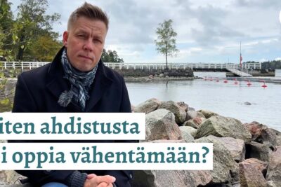 Man sitting on a rocky shoreline near a harbor with Finnish overlay text about learning to reduce anxiety auttaa vahentämään 1.