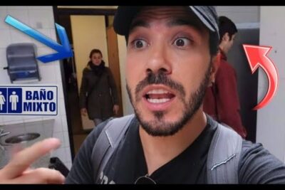 Man in a cap pointing toward a restroom entrance with a visible banos mixtos 1 sign in a tiled public bathroom