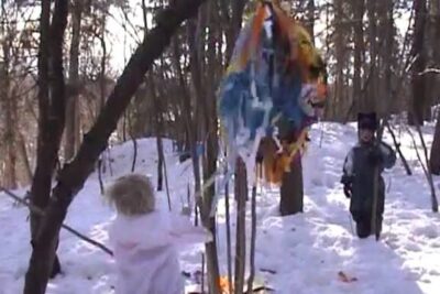 Two children in winter costumes hitting a colorful piñata in the snow at barnehage 2.