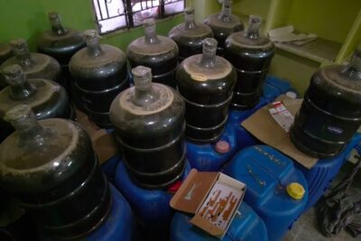 Dust-covered glass carboys stacked on blue plastic drums in a small room for bekampelse narkotikahandel
