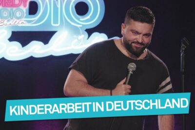 Comedian on stage with microphone and neon sign next to a banner reading Kinderarbeit in Deutschland, ber kinderarbeit 1.