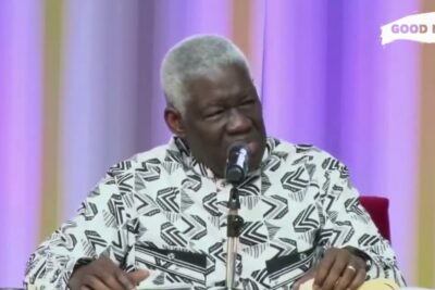Elderly man speaking into a microphone at a table wearing a black-and-white patterned shirt, biens materiels 1.