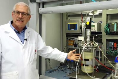 Person in a white lab coat pointing to a pilot bioreactor and control panels for bioteknologisten prosessien