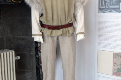 Mannequin wearing a beige medieval linen tunic with fur-trimmed shoulders, belted waist and linen trousers, brugen af 1