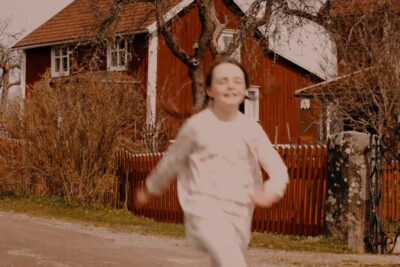 Child running past a red bullerby barn cottage with white trim and wooden fence.