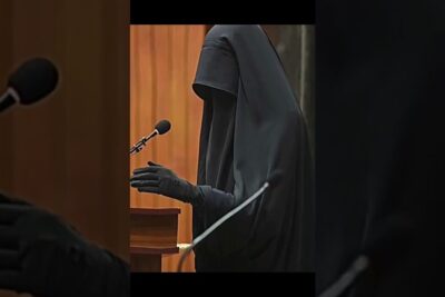 Person wearing a black full-coverage burka ar speaking at a podium with a microphone