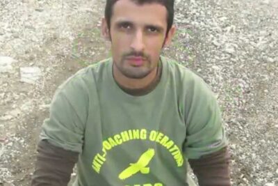 Man seated on gravel wearing a green anti-poaching shirt with a bird logo to draw attention to caccia illegale.