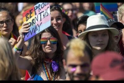 Crowd at a Pride march with rainbow sunglasses, flags and a protest sign, celebrating casamento homossexual 6.