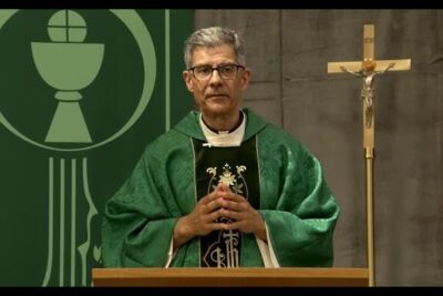 Priest in green vestments leading a catholic church Mass from a wooden lectern with a crucifix nearby.