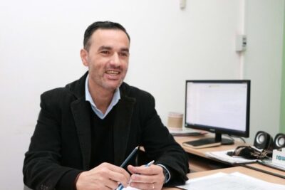 Smiling man at an office desk holding pens with a computer monitor, paperwork and headphones, comercio abierto 1