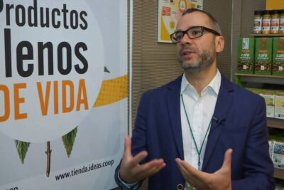 Speaker in a suit gestures at an organic product display and banner promoting comercio justo 2.